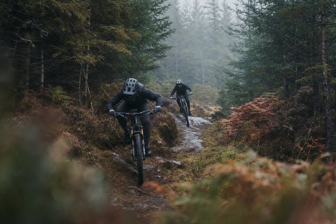 Two mountain bikers descending a rocky forest singletrack between conifers in mid-Wales, shown in black and white